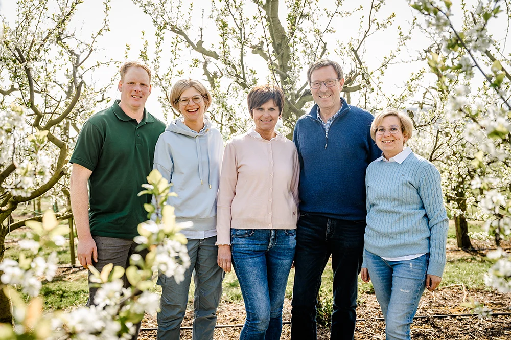 Die Familie Wollseifen steht eng beieinander und lächelnd in einem sonnenbeschienenen Obstgarten mit blühenden Bäumen. Sie sind leger in Pullover und Jeans gekleidet, umgeben von Grünzeug und weißen Blumen und genießen die helle Umgebung.
